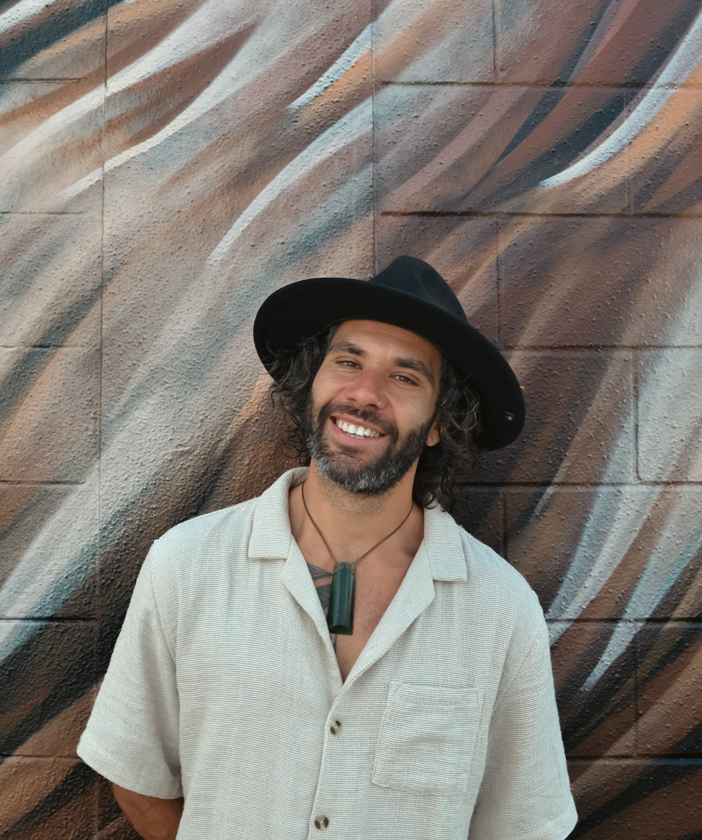 Man wearing a black hat and beige shirt standing against a textured wall.